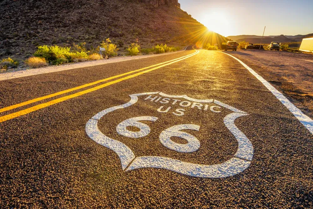 https://www.theamericanroadtripcompany.co.uk/wp-content/uploads/2023/09/Route-66-Street-sign-on-historic-route-66-in-the-Mojave-desert-photographed-against-the-sun-at-sunset.webp